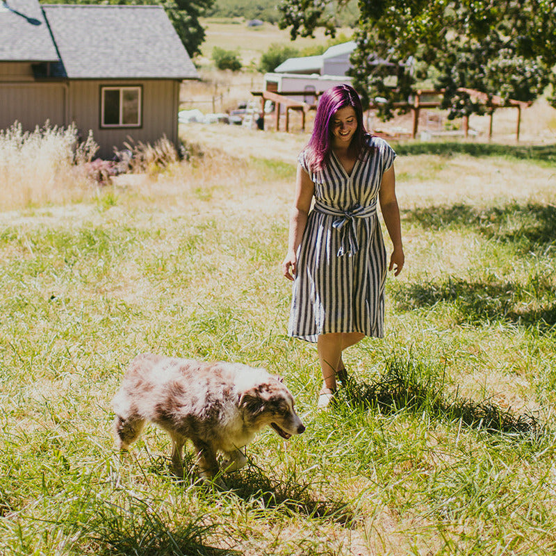Woman with purple hair and a striped dress walking a dog in a grassy field.