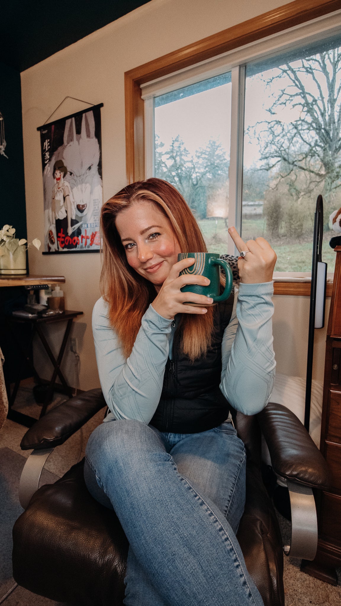 A woman with long red hair sits in a dark leather chair in a home interior, holding a green mug and pointing with her other hand. She is wearing a light blue long-sleeve shirt, a black vest, and blue jeans.