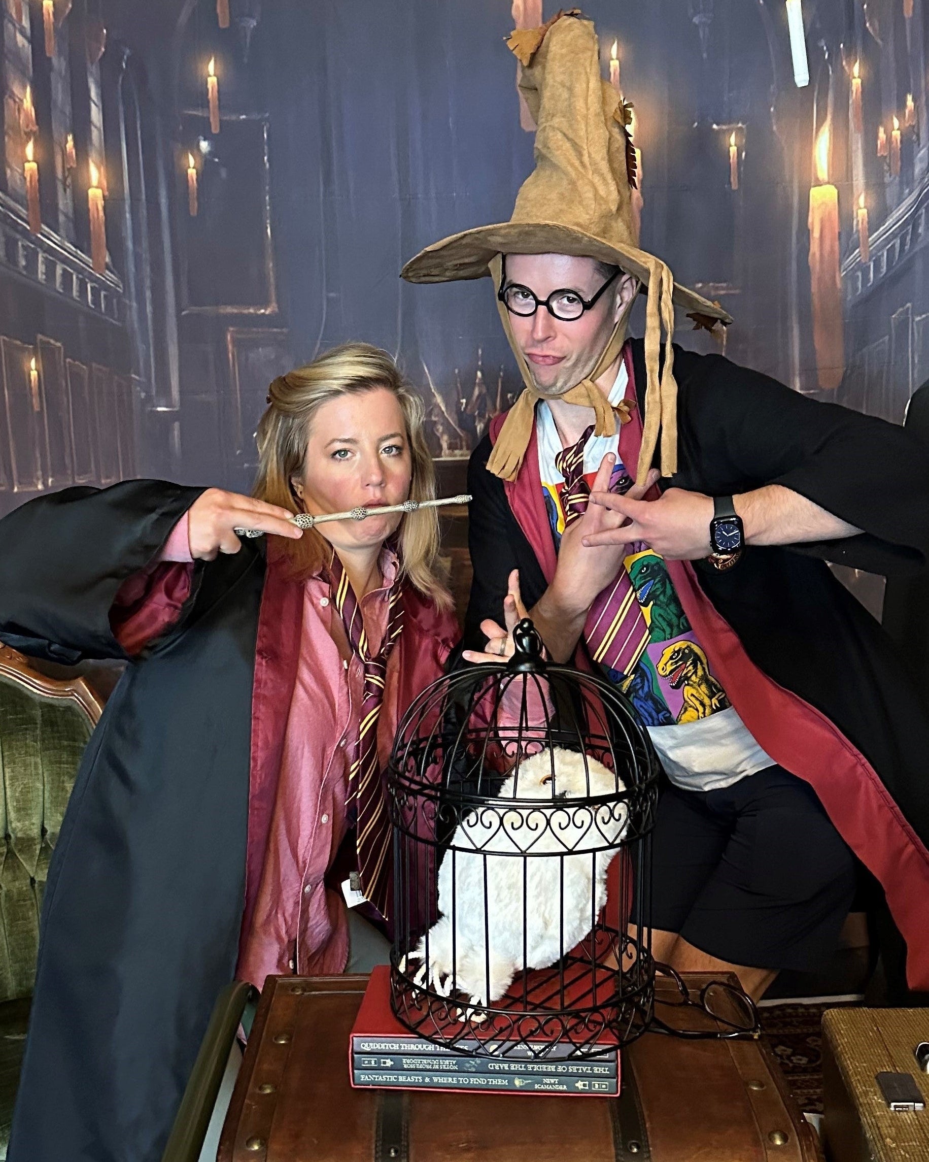 Two people posing in elaborate Harry Potter costumes, including a large witch hat and robes, standing behind vintage suitcases in front of a Hogwarts-themed backdrop with hanging candles.